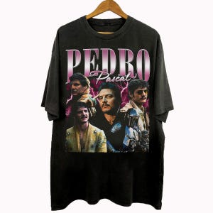 May include: Black t-shirt featuring a graphic of Pedro Pascal with the text "PEDRO Pascal" in a pink and white retro design. The shirt displays multiple images of the actor. The t-shirt is hanging on a wooden hanger.