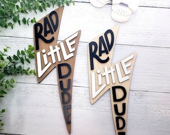 May include: Two wooden lightning bolt shaped signs with the words "Rad Little Dude" in black and white lettering.