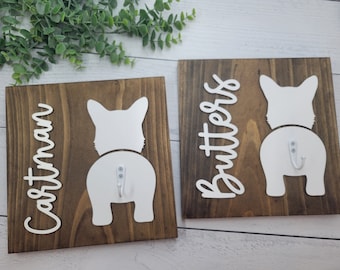 May include: Two wooden signs with white dog silhouettes and hooks. The signs are personalized with the names "Castman" and "Butters".