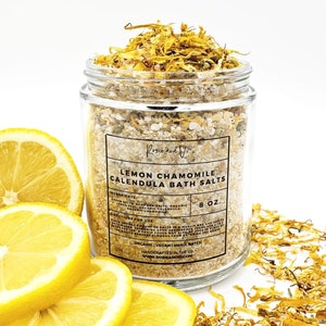 May include: A glass jar filled with bath salts, labeled "Lemon Chamomile Calendula Bath Salts" with a list of ingredients and directions for use. The jar is surrounded by dried yellow flower petals and lemon slices.