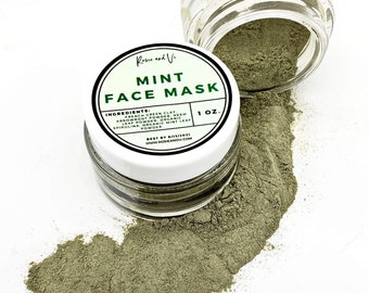 May include: A small glass jar with a white lid containing a green powder labeled "Mint Face Mask". The jar is open and the powder is spilling out onto a white surface. The label also lists the ingredients: French Green Clay, Arbowboot Powder, Neem Leaf Powder, Organic Spirulina, Organic Mint Leaf Powder. The label also states "1 oz." and "Best by 8/13/2021".