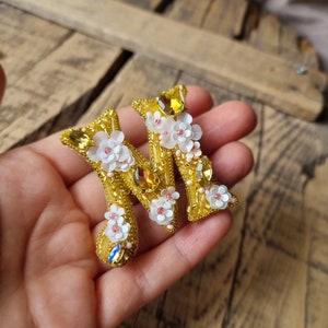 Może przedstawiać: Złota litera M ozdobiona białymi i różowymi kwiatami i żółtymi kryształkami.