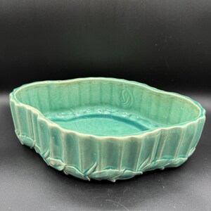 May include: A teal-colored ceramic planter with a ribbed exterior and leaf-like accents. The oval-shaped bowl has a smooth interior and is suitable for small plants or decorative purposes. The planter is set against a dark background.