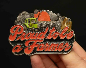 Op de afbeelding: Een zilveren gesp met een rood, groen en zwart emaille ontwerp. De gesp toont een tractor, een schuur en de woorden "Trots om een boer te zijn".