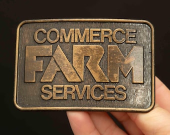 May include: A gold-toned metal belt buckle with the text "COMMERCE FARM SERVICES" embossed on it.