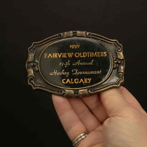 May include: Vintage oval belt buckle with the text "1992 Fairview Oldtimers 17th Annual Hockey Tournament Calgary" in gold lettering. The buckle has a dark background and a decorative bronze-coloured frame.