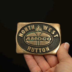 Puede incluir: Una hebilla de cinturón rectangular de latón con las palabras "NORTH WEST AMOCO HUTTON" en un diseño circular. La hebilla tiene un fondo oscuro con un símbolo de antorcha en el centro.