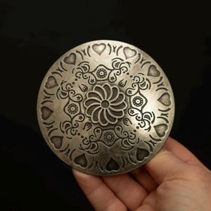 Puede incluir: Un objeto decorativo circular plateado con un intrincado diseño floral y de corazón. El motivo central es una flor estilizada, rodeada de patrones arremolinados y formas de corazón. El objeto se sostiene en una mano sobre un fondo negro.