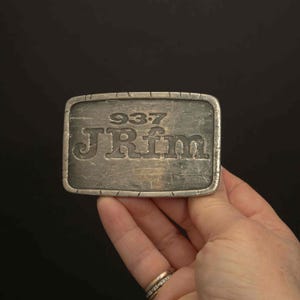 May include: A rectangular silver-toned metal belt buckle with the engraved text "937 JRfm". The buckle has a textured surface and a decorative border. The buckle is held in a hand against a black background.