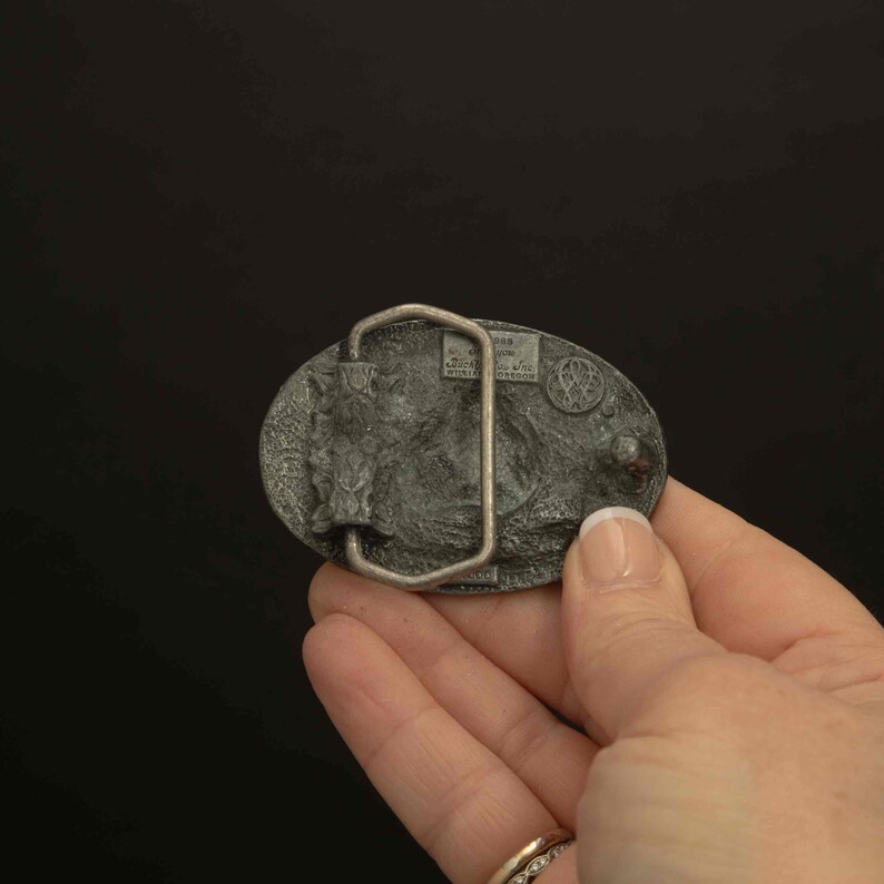 K&ouml;nnte beinhalten: G&uuml;rtelschnalle aus silberfarbenem Metall mit ovaler Form. Die Schnalle zeigt ein detailliertes Reliefdesign eines Pferdes und Text. Die R&uuml;ckseite der Schnalle ist sichtbar und zeigt die G&uuml;rtelschlaufe und die Markierungen des Herstellers. Die Schnalle wird in einer Hand gehalten.