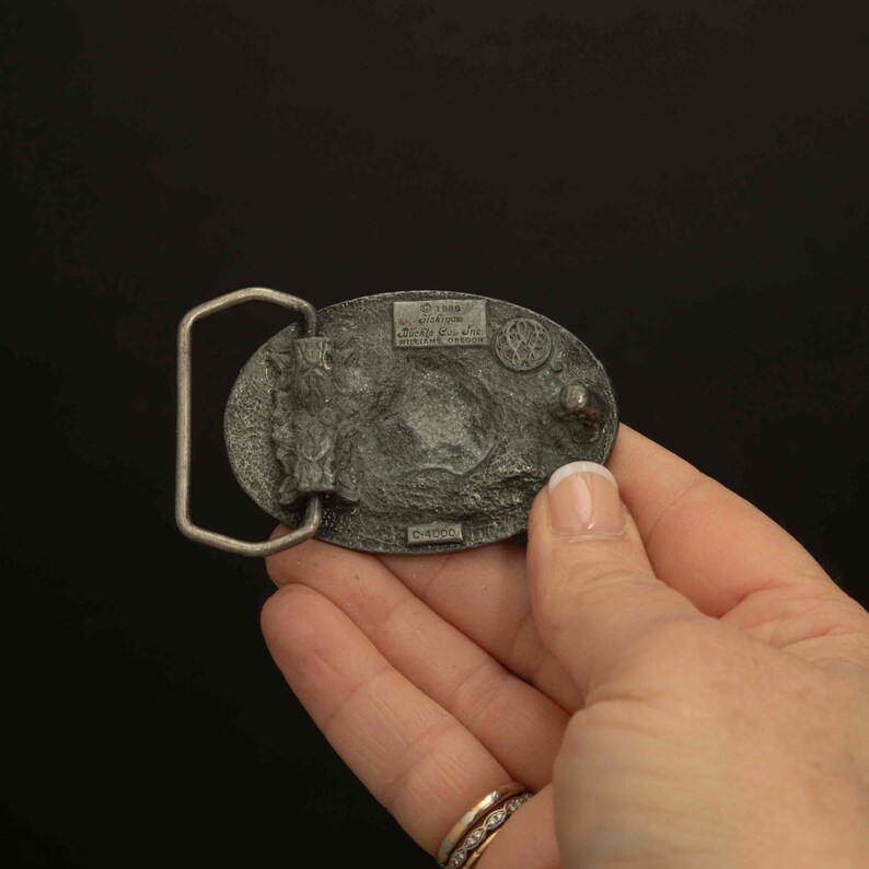 K&ouml;nnte beinhalten: Ovale Metallschnalle mit detailliertem Reliefdesign. Die Schnalle zeigt eine Landschaftsszene mit einem Berg und einem Baum. Der Text "&copy; 1988" und "WILLIAMS, OREGON" ist sichtbar. Die Schnalle wird von einer Hand gehalten.