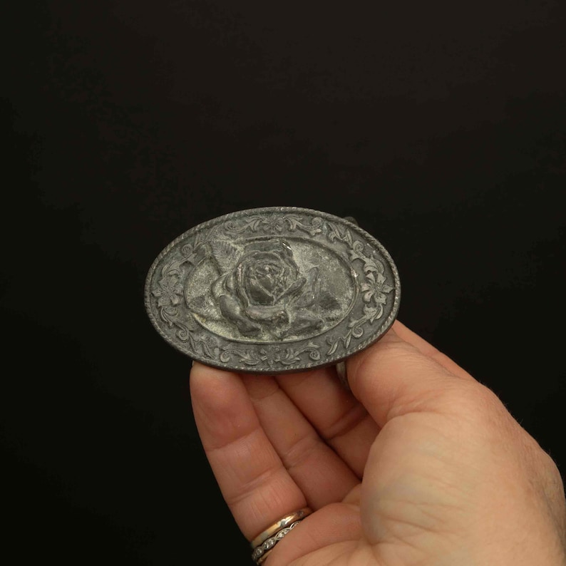 K&ouml;nnte beinhalten: Ovale, silberfarbene G&uuml;rtelschnalle mit detailliertem Rosendesign in der Mitte. Die Schnalle hat einen verzierten Rand mit Blumen- und Bl&auml;ttermustern. Die Schnalle wird in einer Hand vor schwarzem Hintergrund gehalten.