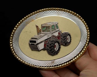 Puede incluir: Una hebilla de cinturón dorada con un tractor blanco con detalles rojos y negros. La hebilla tiene un óvalo plateado con un patrón de diamante.