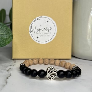 Può includere: Un braccialetto con perline nere e legno con un ciondolo a forma di loto argentato. Il braccialetto è confezionato in una piccola scatola con un adesivo che dice "Lulaverse Hand Made Jewelry".