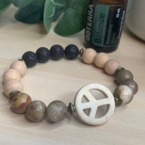 Pode incluir: Um bracelete de contas com um amuleto de sinal de paz. O bracelete é feito com contas marrons, bege e pretas. O amuleto de sinal de paz é branco com um contorno marrom.