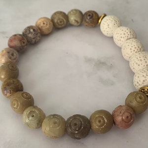 Op de afbeelding: Een armband gemaakt van bruine, beige en witte kralen. De kralen zijn rond en hebben een getextureerd oppervlak. De armband heeft een gouden sluiting.