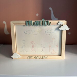 May include: A wooden picture frame with a child's drawing of a family and a house. The frame has the name "marin" written on it in green letters and the words "art gallery" written on the bottom.