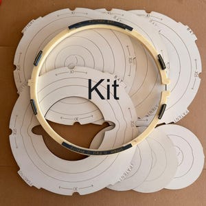 Pode incluir: Um kit de pratos para torno de oleiro com vários discos brancos circulares com marcas impressas e o texto 30 e 30,5. Um anel grande de cor branco-sujo com clipes escuros e o texto Pottery Bat, Simple, Fast, Precise está sobre os discos.
