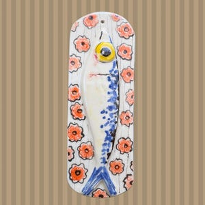 Puede incluir: Un colgante de pared con forma de pez de cerámica. El pez es blanco con manchas azules y una cola azul. Tiene un ojo amarillo grande. El fondo es blanco con diseños de flores naranjas. La pieza mide unos 25 cm de largo.