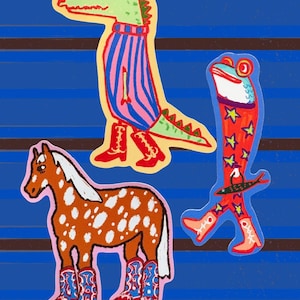 May include: Three stickers featuring a green alligator in a pink and blue striped shirt and red boots, a frog in red star-patterned trousers and red boots holding a fish, and a brown and white spotted horse wearing blue and red cowboy boots.