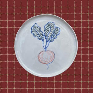 May include: A white ceramic plate featuring a hand-drawn radish design. The radish has a red root, blue stem, and green leaves. The plate is round and rests on a red checkered tablecloth.