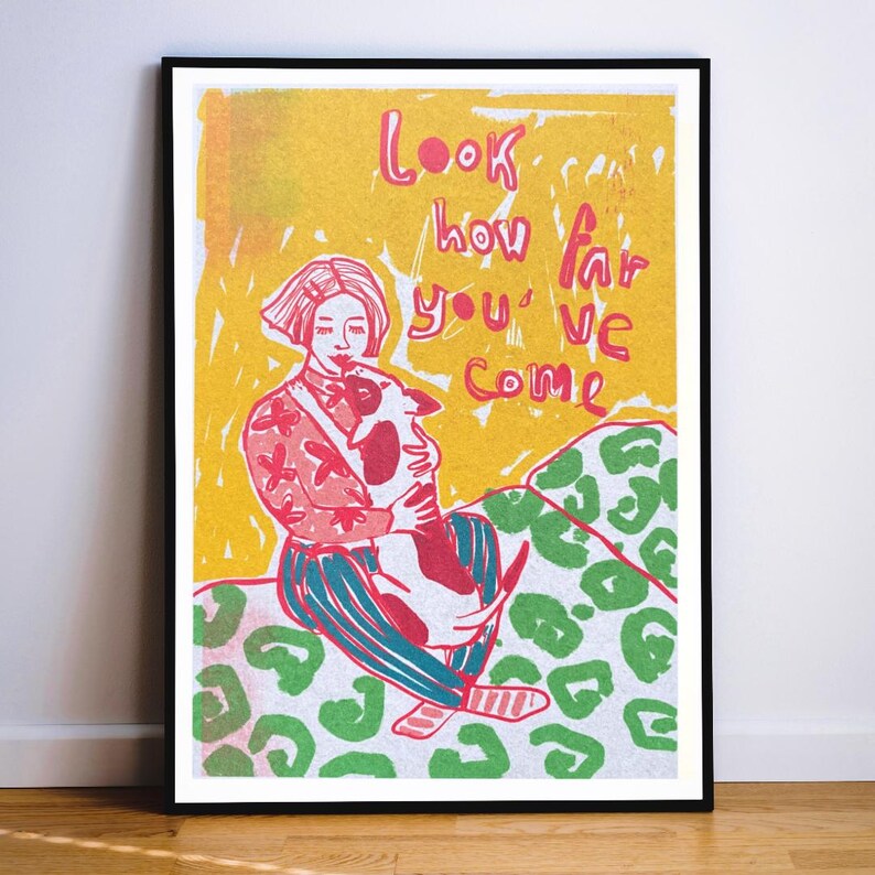 May include: A framed art print featuring a person with short hair holding a small dog. The person wears a pink patterned top and blue striped pants. The background is yellow with green leopard spots. The text reads: Look how far you've come.