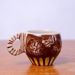May include: A brown ceramic mug with a white floral design and a yellow and white striped base. The mug has a unique, twisted handle.