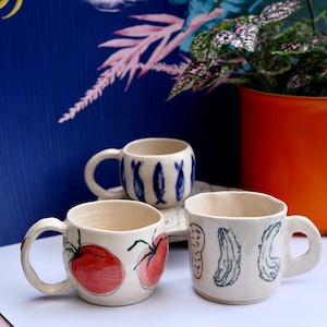 Könnte beinhalten: Drei handgefertigte Keramikbecher mit einzigartigen Designs. Ein Becher zeigt rote Tomaten, ein anderer blaue Fische und der dritte grüne Gemüse. Jeder Becher hat einen weißen Hintergrund und einen abgerundeten Griff. Die Becher stehen auf einer weißen Oberfläche mit blauem Hintergrund.