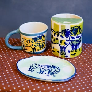 May include: A ceramic set consisting of a mug with a blue handle, a cylindrical container, and an oval dish. Each piece features hand-painted illustrations of sheep in blue and yellow against a light green and white checkered or spotted background.