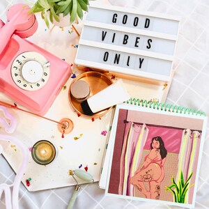 K&ouml;nnte beinhalten: Ein rosa Drehwahltelefon, eine wei&szlig;e Lichtbox mit dem Text "GOOD VIBES ONLY", eine kleine Kerze, eine Jade-Walze, eine Tube Lotion, ein kleines Glas Creme und ein Skizzenbuch mit einer Zeichnung einer Frau in einem rosa Badeanzug, die in einem Badezimmer mit einer gr&uuml;nen Pflanze steht.
