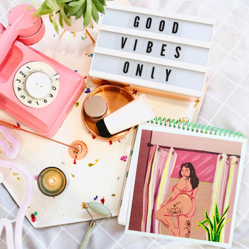 K&ouml;nnte beinhalten: Ein rosa Drehwahltelefon, eine wei&szlig;e Lichtbox mit dem Text "GOOD VIBES ONLY", eine kleine Kerze, eine Jade-Walze, eine Tube Lotion, ein kleines Glas Creme und ein Skizzenbuch mit einer Zeichnung einer Frau in einem rosa Badeanzug, die in einem Badezimmer mit einer gr&uuml;nen Pflanze steht.