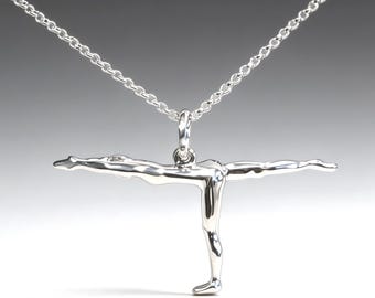 May include: A silver pendant necklace showing a yoga pose. The pendant depicts a person in the Warrior III pose, with arms and one leg extended. The necklace chain is a delicate silver link.