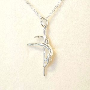 Può includere: Una collana con pendente color argento raffigurante una ballerina su un palo. La ballerina è in una posa aggraziata, con le braccia tese. Il pendente è appeso a una delicata catena d'argento. Lo sfondo è beige chiaro.