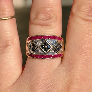 Puede incluir: Un anillo dorado con una banda de pequeñas gemas rojas vibrantes. El centro presenta un patrón geométrico de gemas negras y blancas. El anillo se lleva en un dedo, mostrando su intrincado diseño y artesanía.