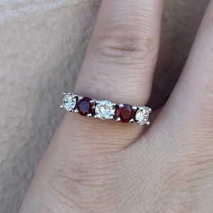 Puede incluir: Un anillo plateado con piedras preciosas redondas y transparentes alternadas con piedras preciosas de color rojo intenso. El anillo se lleva en un dedo, mostrando el contraste entre las piedras brillantes y el tono de la piel. El diseño del anillo es clásico y elegante.