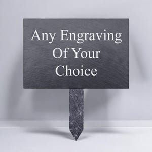 May include: A rectangular black slate garden marker with a pointed stake at the bottom. The surface features the white engraved text Any Engraving Of Your Choice centred in three lines with a serif font style against the dark stone texture.