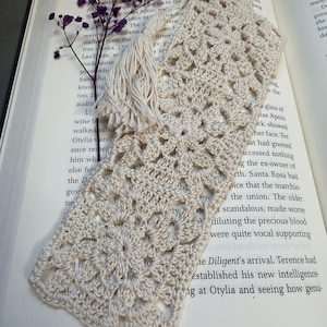 May include: A rectangular off-white crochet bookmark featuring a repeating floral motif pattern and a matching tassel attached to the top. A small sprig of dried purple flowers is placed alongside the bookmark on an open book page.