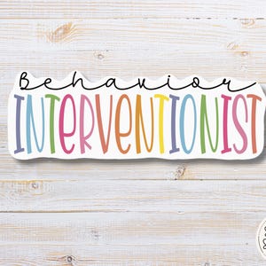 May include: A colorful sticker with the words "Behavior Interventionist" in a playful font. The word "Behavior" is in black script above the word "Interventionist", which is in rainbow colors. The sticker is on a white wooden background.