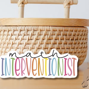 May include: A colorful sticker with the text "math interventionist" in a rainbow of colors. The sticker is on a wooden surface.