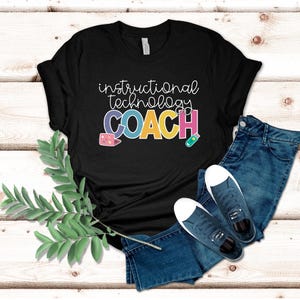 May include: Black t-shirt with the words "instructional technology COACH" in a colorful design. The shirt is paired with blue jeans and navy blue sneakers. A sprig of green leaves is on the left side of the image.