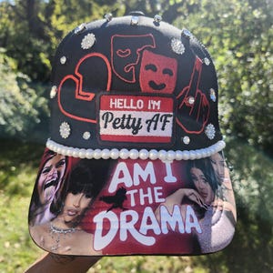 May include: Black baseball cap with a curved brim, embellished with rhinestones and pearls. The cap features the text "HELLO I'M Petty AF" and "AM I THE DRAMA" with images of women. The design includes red and white accents.