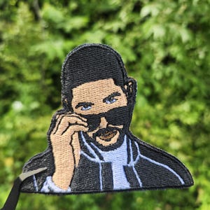 May include: Embroidered patch depicting a person wearing sunglasses. The patch has a black outline, with tan skin tones, black hair, and blue accents. The person is holding up their sunglasses. The patch is held by tweezers against a green background.