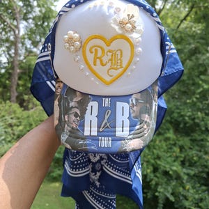 May include: A white trucker hat featuring a yellow embroidered heart with the letters RB, adorned with white floral and pearl embellishments. The brim displays a graphic print with the text THE R&B TOUR, and a blue bandana is attached to the sides.