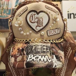 Puede incluir: Una gorra de béisbol beige con un parche marrón en forma de corazón bordado con iniciales. La gorra presenta detalles de perlas, adornos florales y el texto "RUN IT". También se ve una cadena dorada y una tela estampada con el texto "BREEZY BOWL".