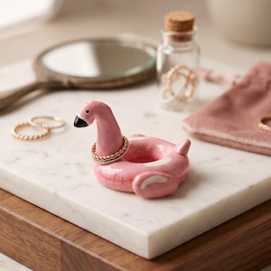 May include: A pink ceramic flamingo ring holder shaped like an inflatable pool float with a black and white beak. The flamingo neck serves as a post for holding rings, and the base is a circular ring-shaped float with painted white accents.