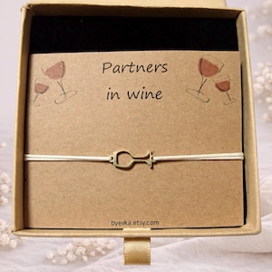 Puede incluir: Un brazalete con un dije de copa de vino dorado, presentado en una caja de regalo. La tarjeta interior dice "Partners in wine" con ilustraciones de copas de vino. La URL de la tienda Etsy es visible.