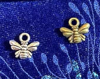 Puede incluir: Dos pequeños dijes de abeja. Uno es plateado y el otro dorado. Ambos dijes tienen un lazo en la parte superior para sujetarlos a la joyería. Las abejas están sobre una superficie azul texturizada con un lápiz en el fondo.