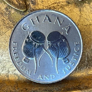 May include: A silver coin with the words "GHANA FREEDOM AND JUSTICE" around the edge. The center of the coin features two drums with a design of a traditional African drum.