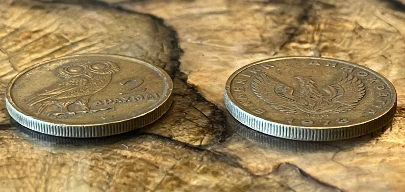 May include: Two vintage Greek coins. One coin features an owl and the text "2 DRAXMAI". The other coin displays an eagle and the text "&Epsilon;&Lambda;&Lambda;&Eta;&Nu;&Iota;&Kappa;&Eta; &Delta;&Eta;&Mu;&Omicron;&Kappa;&Rho;&Alpha;&Tau;&Iota;&Alpha; 1974". Both coins are bronze-colored and have a ridged edge.