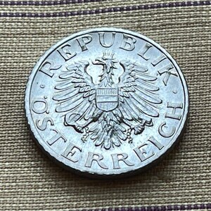 May include: A silver Austrian coin with the inscription "REPUBLIK ÖSTERREICH" and a depiction of the Austrian coat of arms featuring an eagle with a shield.